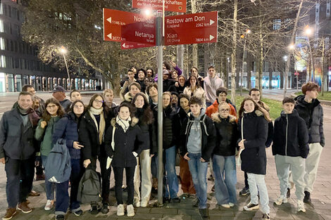 Around 35 people stand in the evening darkness on a brightly lit central island of a metropolitan street at a signpost pointing in the direction of several cities. The top sign reads "Partnerships of the City of Dortmund".