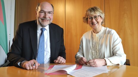 Two people are sitting next to each other at a table on which papers are lying.