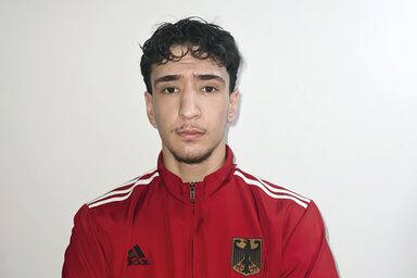 A man in a red tracksuit jacket with the German coat of arms on his chest looks into the camera with his arms folded.