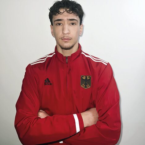 A man in a red tracksuit jacket with the German coat of arms on his chest looks into the camera with his arms folded.