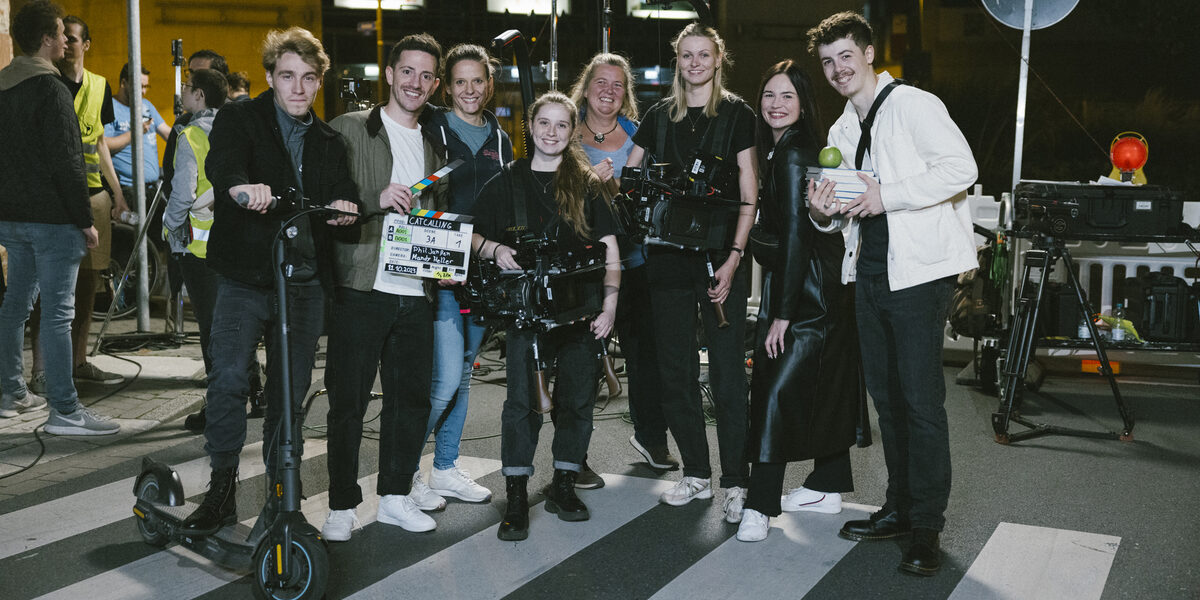 A group of people are standing on a crosswalk, one person is holding an e-scooter, two other people are holding a film camera and a film clapperboard.