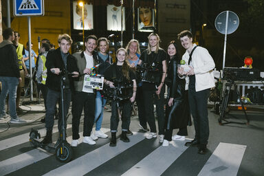 Eine Gruppe von Personen steht auf einem Zebrastreifen, eine Person hält einen E-Scooter, zwei andere Personen halten eine Filmkamera und eine Filmklappe.