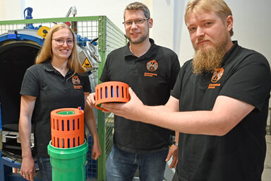 Drei Personen stehen im Hochspannungslabor der FH Dortmund und zeigen ein Bauteil eines Batteriespeichers.