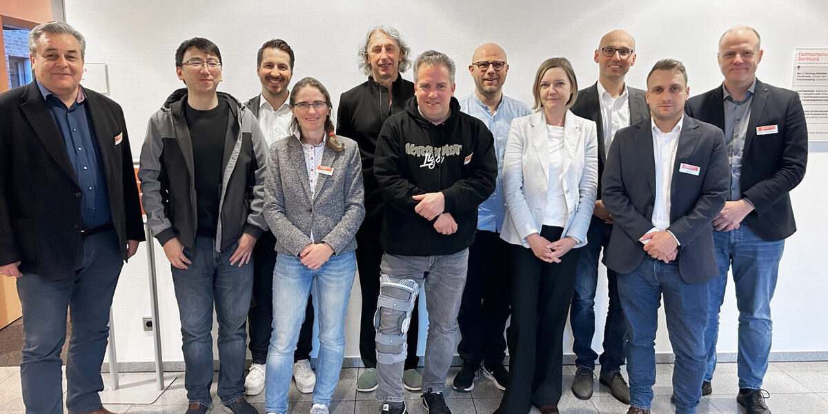 Elf Personen stehen nebeneinander vor einer hellen Wand und schauen freundlich in die Kamera.