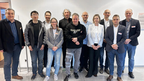 Elf Personen stehen nebeneinander vor einer hellen Wand und schauen freundlich in die Kamera.