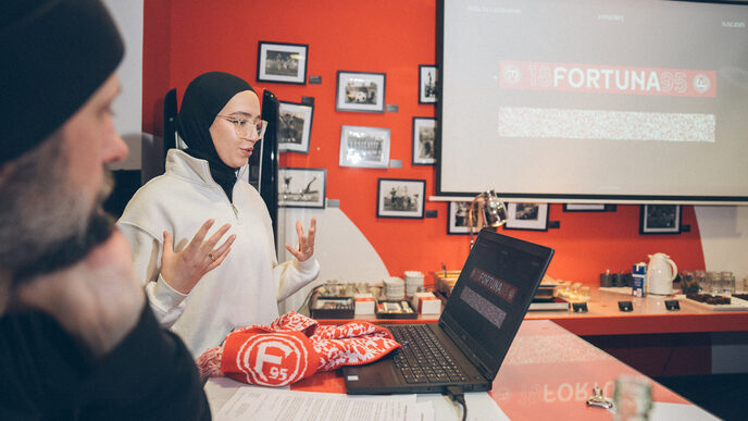 A female person reading explains something while looking at a laptop. The design for a fan scarf can be seen in the background.
