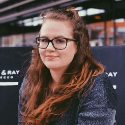 Gastwissenschaftlerin Dr. Charlotte Rottiers am Fachbereich Architektur. Sie trägt einen dunklen Pullover und lächelt leicht in die Kamera. Im Hintergrund sind unscharfe Schilder und städtische Strukturen zu sehen.