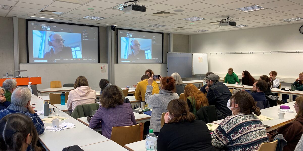 Menschen sitzen in einem Seminarraum und blicken auf die Projektion eines Video-Calls.