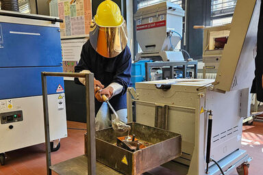 One person wears a protective helmet with a visor and holds a ladle from which his silvery substance flows into a mold.