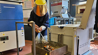 One person wears a protective helmet with a visor and holds a ladle from which his silvery substance flows into a mold.