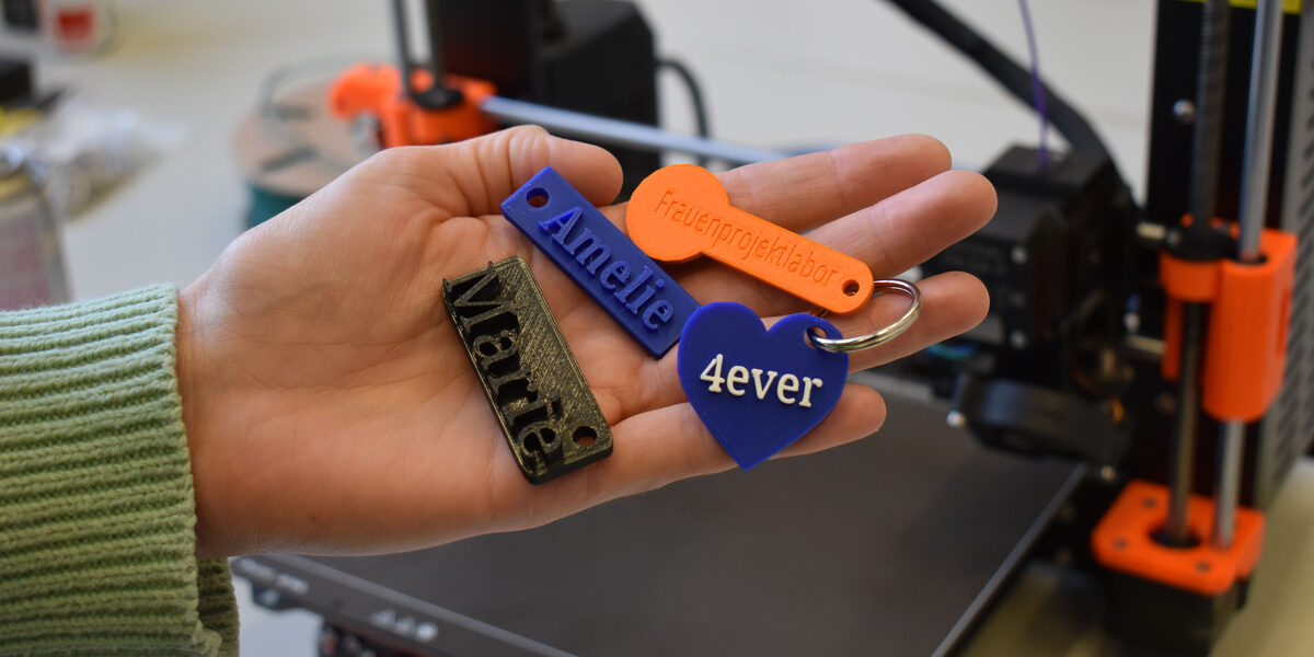 Four differently designed key rings lie in the palm of one hand, with a 3D printer in the background.