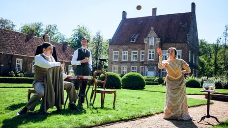 Eine Frau wirft einen Ball vor einem historischen Gebäude. Drei Männer beobachten sie kritisch.