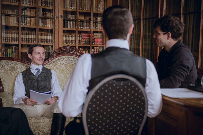 People in period dress from the time of the German Empire sit together and read a script.