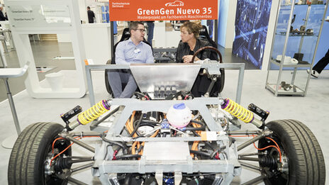 Two people are sitting in a vehicle at a trade fair stand and talking.