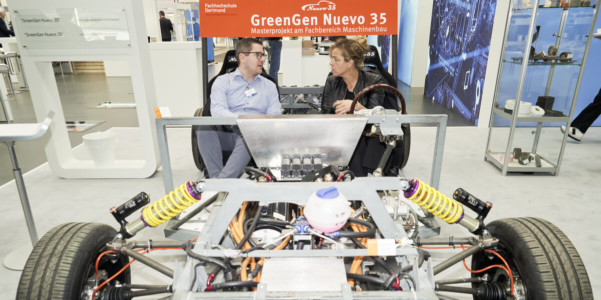 Two people are sitting in a vehicle at a trade fair stand and talking.