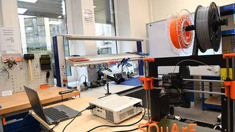 Das Bild zeigt einen 3D-Drucker mit dem Ausgedruckten dreidimensionalen Schiftzug SQuArE, im Hintergrund ist eine Werkbank zu sehen.