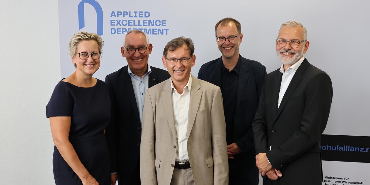 Fünf Personen in repräsentablem Outfit lächeln in die Kamera. Auf dem Banner im Hintergrund ist "Applied Excellence Department" zu lesen.