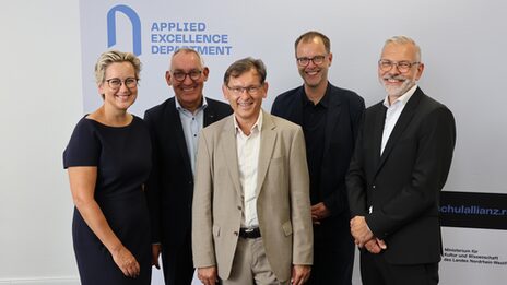Five people in prestigious outfits smile at the camera. The banner in the background reads "Applied Excellence Department".