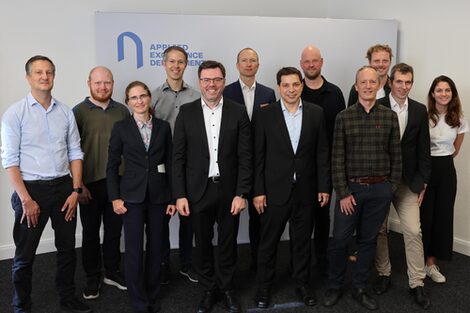 Twelve people pose in front of a stand labeled "Applied Excellence Department" and look smilingly into the camera.