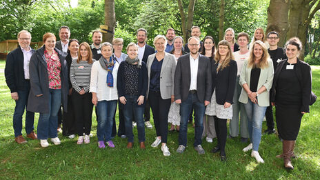 23 Personen stehen auf einer Wiese unter Bäumen und vor Büschen, durch die Baumkronen fällt Sonnenlicht.