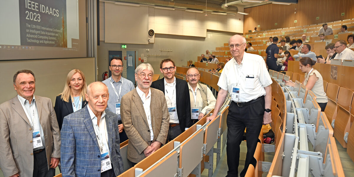 A group of people are standing in a lecture hall. The words "IEEE IDAACS 2023" can be read on the screen.