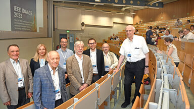 Eine Gruppe Menschen steht in einem Hörsaal. Auf der Leinwand ist der Schriftzug "IEEE IDAACS 2023" zu lesen.