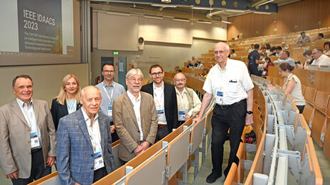 A group of people are standing in a lecture hall. The words "IEEE IDAACS 2023" can be read on the screen.