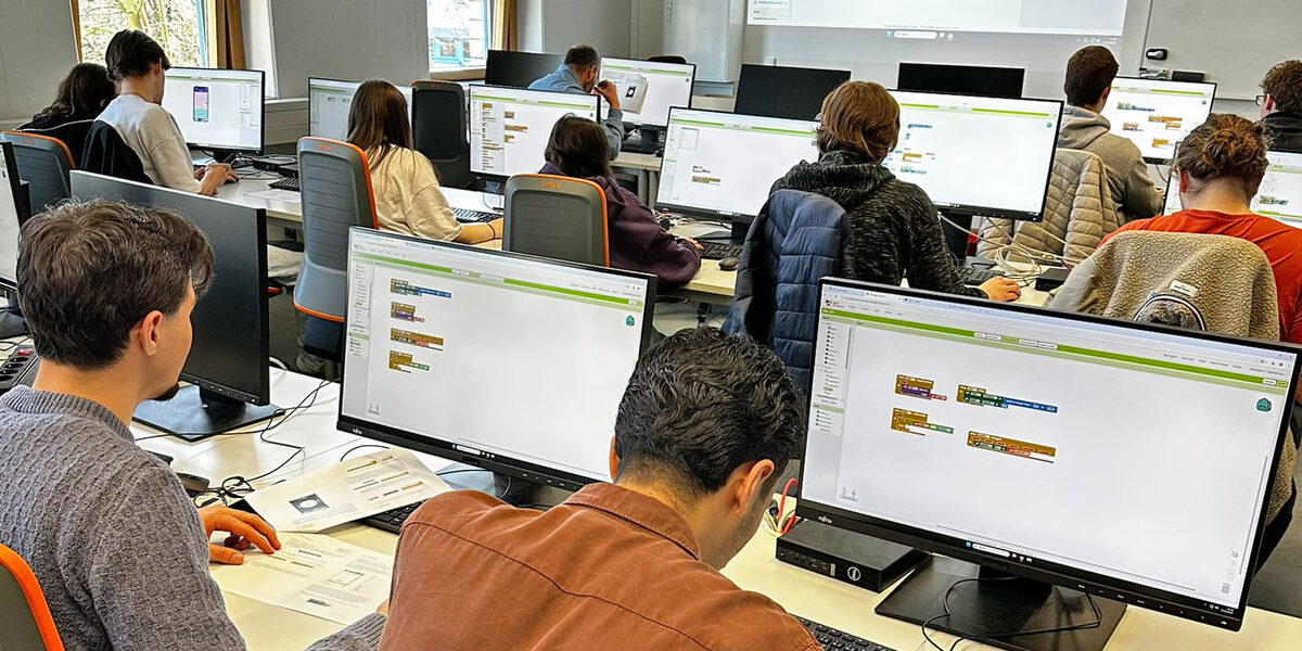 Several people are sitting in a room with computer screens on the tables in front of them.