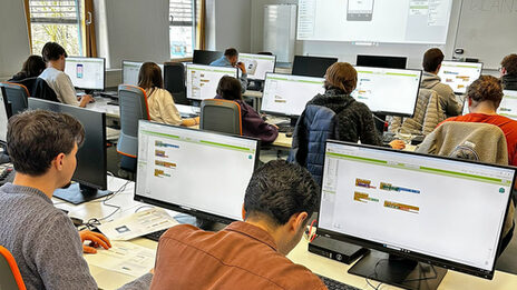 Mehrere Personen sitzen in einem Raum, vor ihnen stehen Computerbildschirme auf den Tischen.