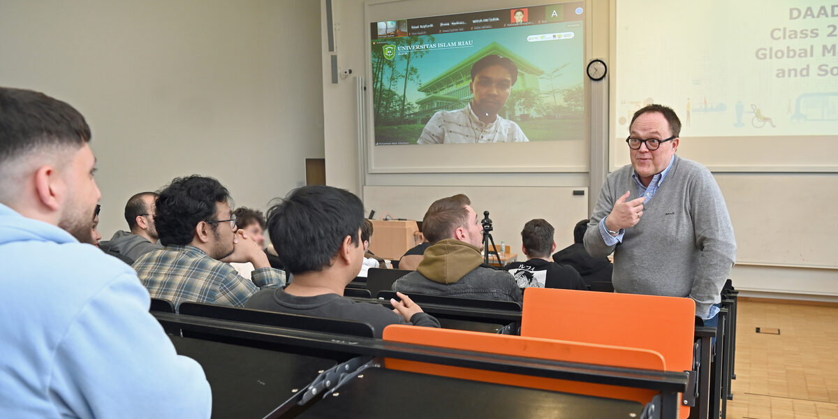 A person read as a male stands in a lecture hall and speaks to a group of students with commitment. Another person is digitally connected on the screen.