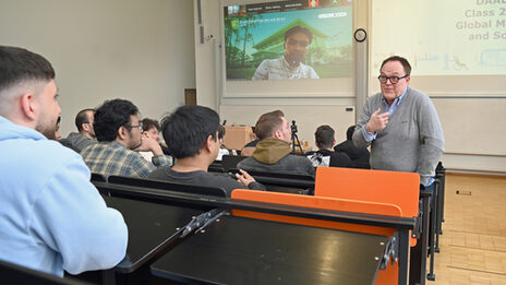 A person read as a male stands in a lecture hall and speaks to a group of students with commitment. Another person is digitally connected on the screen.