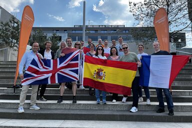 Mehrere Menschen stehen draußen auf einer Treppe, umrahmt von zwei Beachflags der Fachhochschule Dortmund. Die Personen in der ersten Reihe halten vor sich Flaggen der Länder Großbritannien, Spanien und Frankreich.