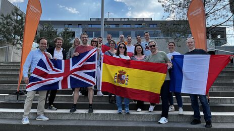 Mehrere Menschen stehen draußen auf einer Treppe, umrahmt von zwei Beachflags der Fachhochschule Dortmund. Die Personen in der ersten Reihe halten vor sich Flaggen der Länder Großbritannien, Spanien und Frankreich.
