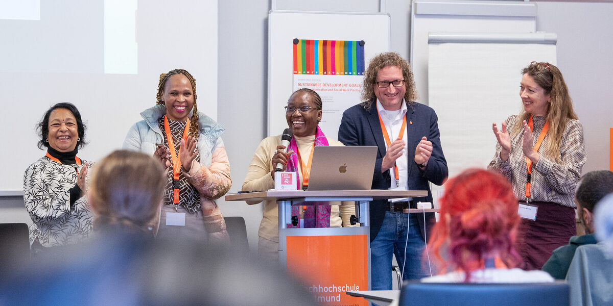 Fünf Personen stehen an einem Redenpult, die mittlere hält ein Mikro, die anderen klatschen, alle lächeln. Im Vordergrund sind die Hinterköpfe sitzender Menschen zu sehen.