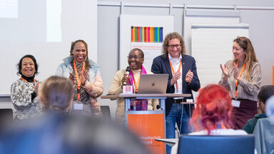 Fünf Personen stehen an einem Redenpult, die mittlere hält ein Mikro, die anderen klatschen, alle lächeln. Im Vordergrund sind die Hinterköpfe sitzender Menschen zu sehen.