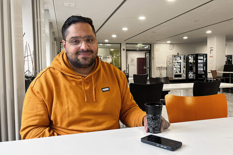 Eine Person im orangefarbenen Kapuzenpullover sitzt an einem Tisch in einem größeren Raum mit mehreren Tischen und Stühlen. Vor ihm steht ein Kaffeebecher, daneben liegt ein Handy.
