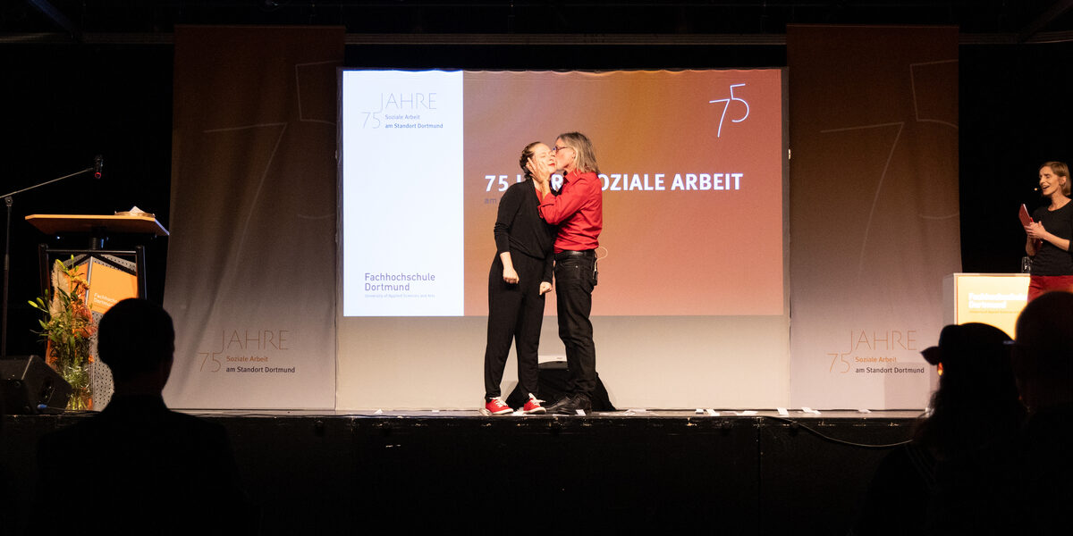 Zwei Personen auf einer Bühne, die rechte gibt der linken einen Kuss auf die Wange. Im Hintergrund ist ein Beamerbild mit dem Text „75 Jahre Soziale Arbeit“ zu sehen.