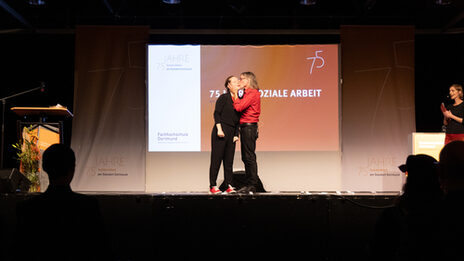 Eindrücke von der FeierZwei Personen auf einer Bühne, die rechte gibt der linken einen Kuss auf die Wange. Im Hintergrund ist ein Beamerbild mit dem Text „75 Jahre Soziale Arbeit“ zu sehen.