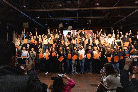 Many people with orange folders in their hands throw their black doctor's hats in the air while being filmed by several people in the foreground.