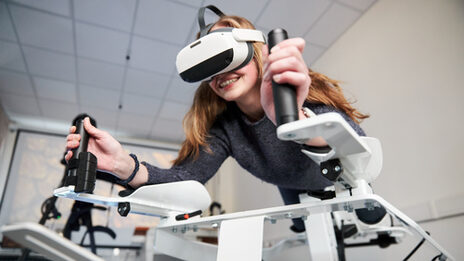 Photo of a person on the Icaros device. She is wearing VR goggles and holding on to the handles.
