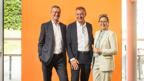 Two men and one woman stand in front of an orange LED wall and smile into the camera.