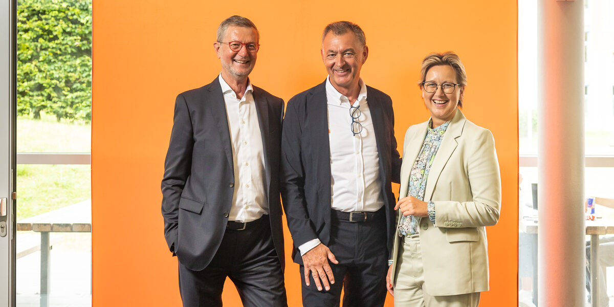 Two men and one woman stand in front of an orange LED wall and smile into the camera.