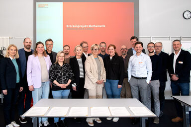 A large group is standing in front of a projector on which the words "Brückenprojekt Mathematik, Feierstunde zum 8. Jubiläum" can be read.