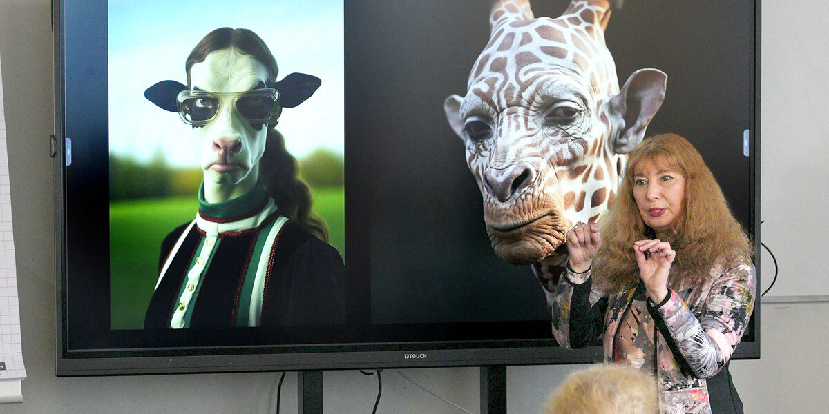 Eine Person spricht und gestikuliert vor einem großen Bildschirm, auf dem zwei offensichtlich künstlich erzeugte Porträts von Mensch-Tier-Mischwesen zu sehen sind.