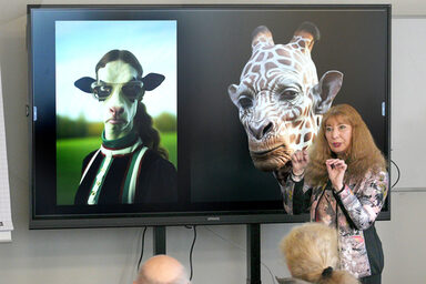 A person speaks and gesticulates in front of a large screen on which two obviously artificially created portraits of human-animal mixtures can be seen.