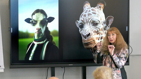 A person speaks and gesticulates in front of a large screen on which two obviously artificially created portraits of human-animal mixtures can be seen.