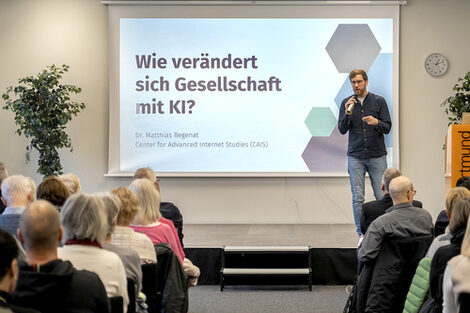 A man stands on a low stage in front of a full row of chairs. He speaks into the microphone in his hand. On the wall behind him is a projector image with the text "How is society changing with AI?" Dr. Matthias Begenat, Center for Advanced Internet Studies.