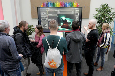 Seven people are standing around a large screen. One of them speaks to the others and points in the direction of the screen. The screen shows the desktop of a video program with the motif of a car.