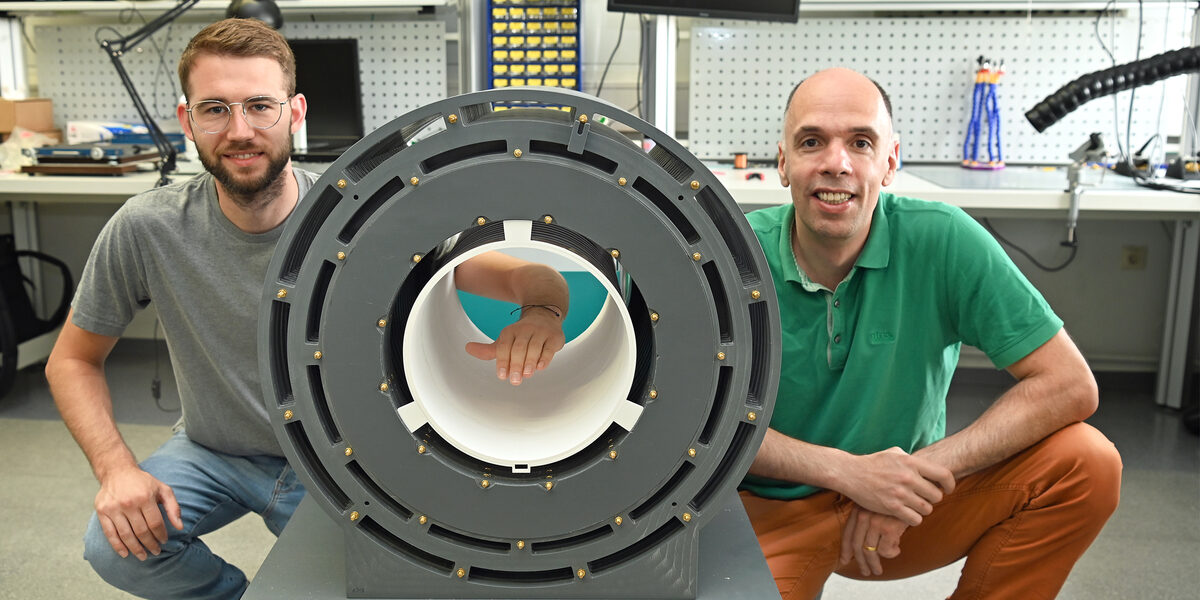 Two people are squatting next to a tube-like construction in a laboratory. One person holds their arm into the tube.