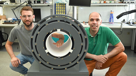 Two people are squatting next to a tube-like construction in a laboratory. One person holds their arm into the tube.